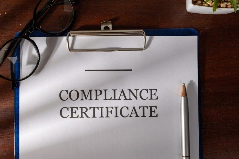 Compliance certificate on clipboard with pen and glasses nearby.