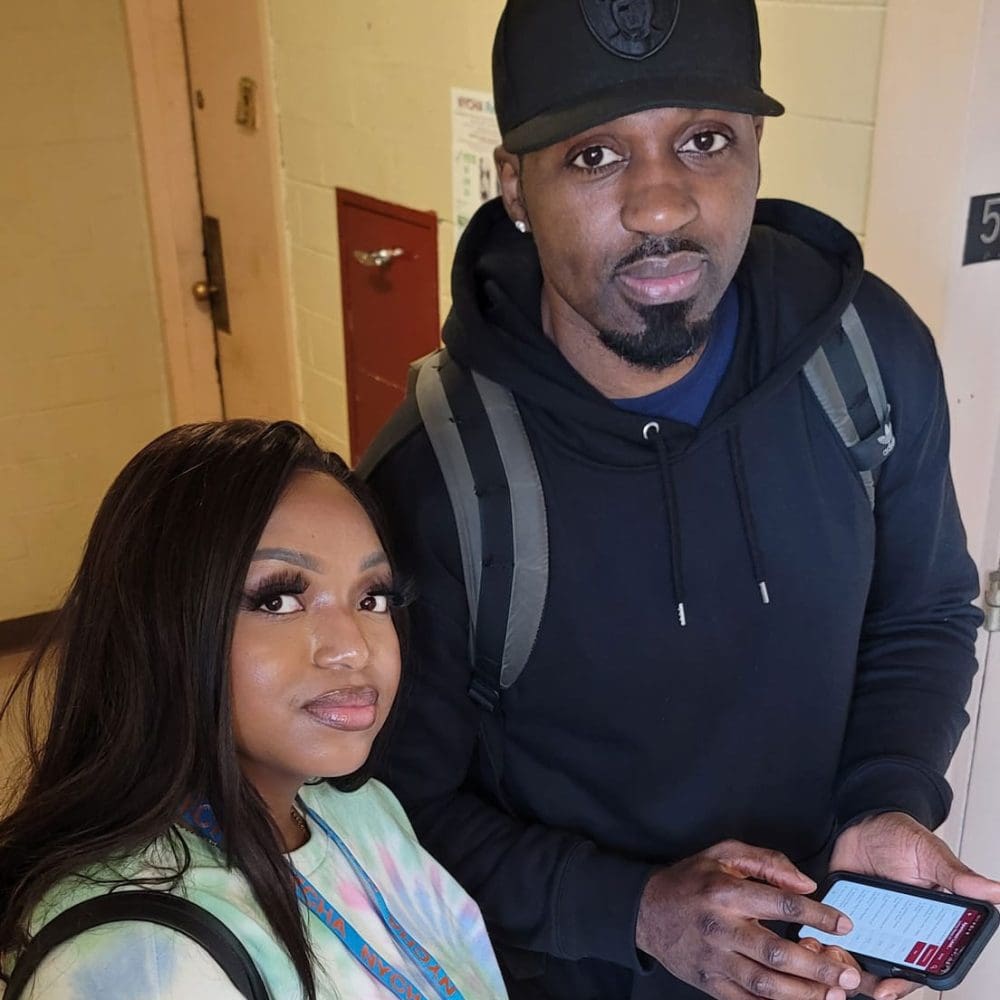 Two people standing indoors, one with phone, both looking at camera.