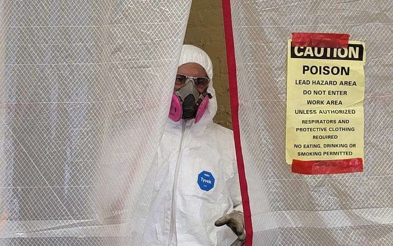 Worker in protective suit near lead hazard warning sign.