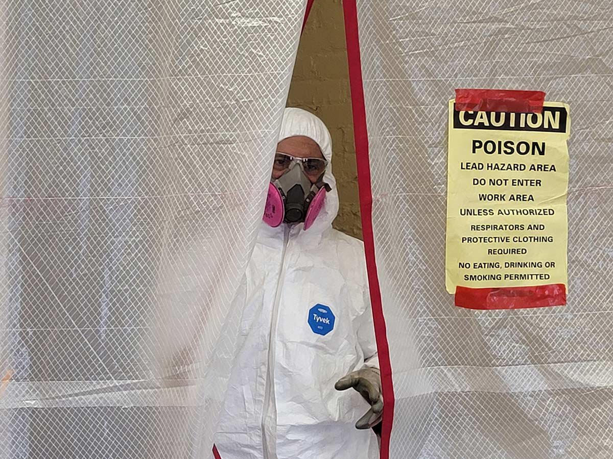 Worker in protective suit with caution sign for lead hazard area.