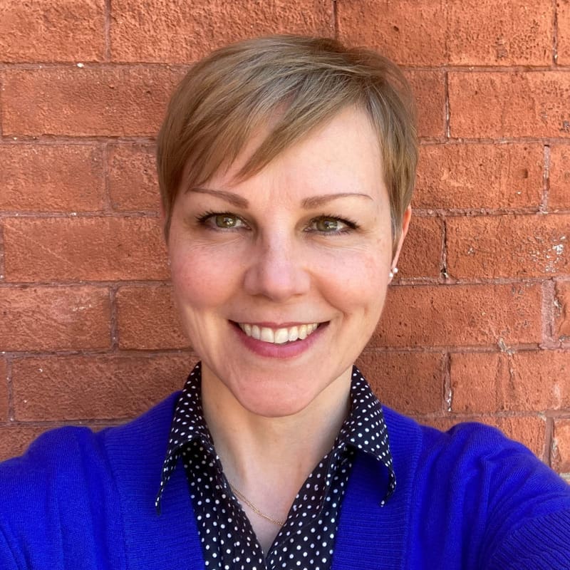 Smiling person with short hair against a brick wall background.
